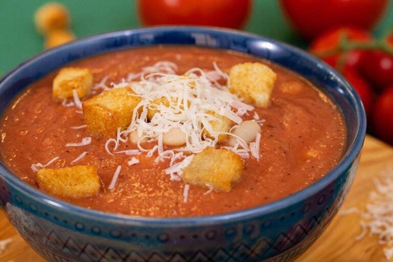 A low side shot of 3-ingredient white bean tomato soup in a blue bowl on a cutting board, topped with croutons and parmesan cheese.