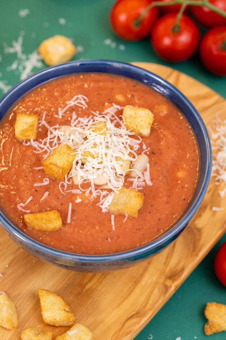 A close up shot of 3-ingredient white bean tomato soup in a blue bowl on a cutting board, topped with croutons and parmesan cheese.
