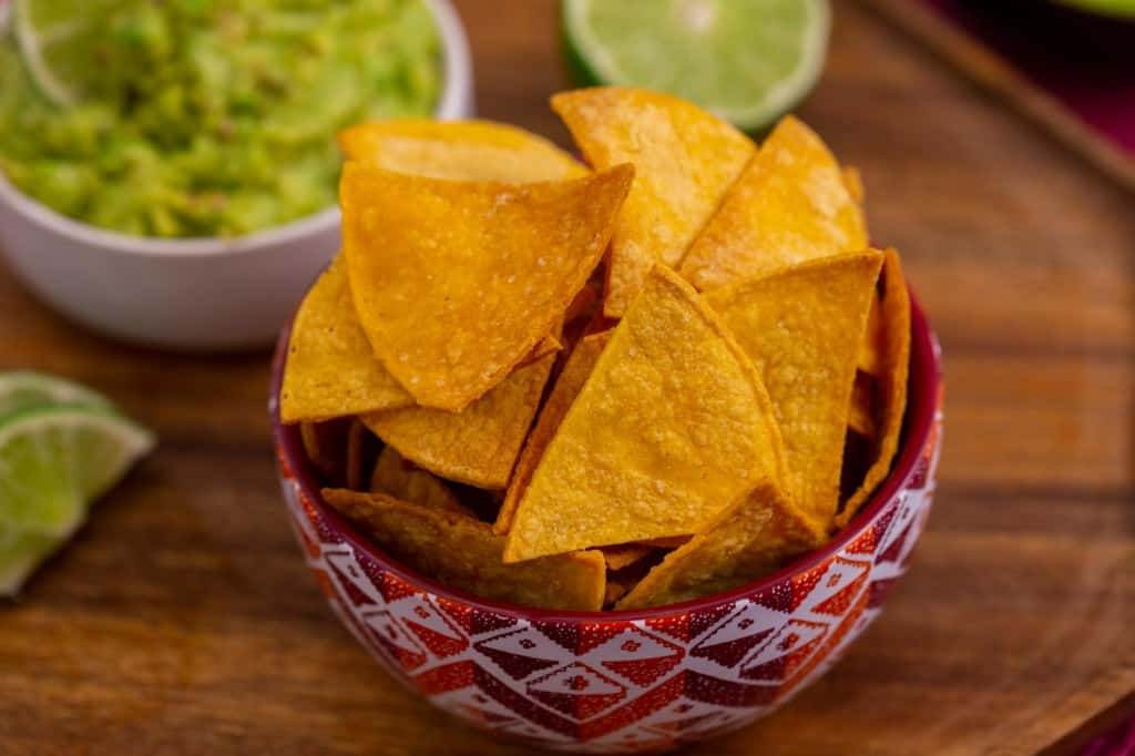 A bowl of air fryer tortilla chips on a wooden cutting board with sliced limes on the side and a bowl of guacamole in the background.