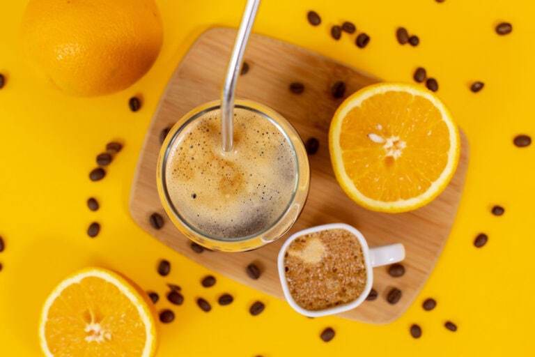 An overhead photo of an iced coconut water americano on a yellow table next to a sliced orange and a small cup of espresso
