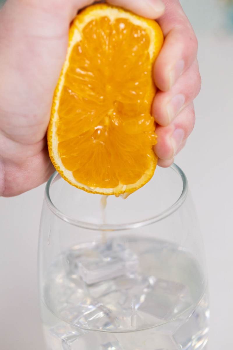 Someone squeezing half of an orange into the glass of iced coconut water
