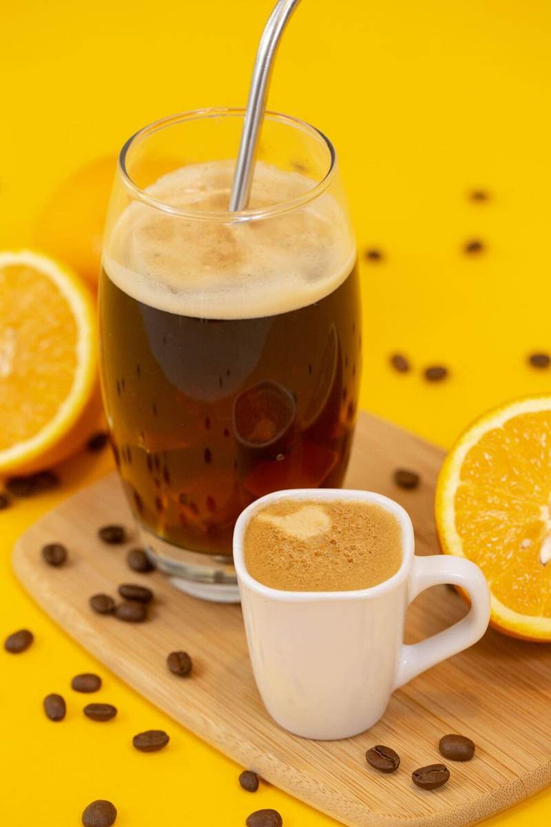 Another photo of the americano on a yellow table next to a sliced orange and a cup of espresso