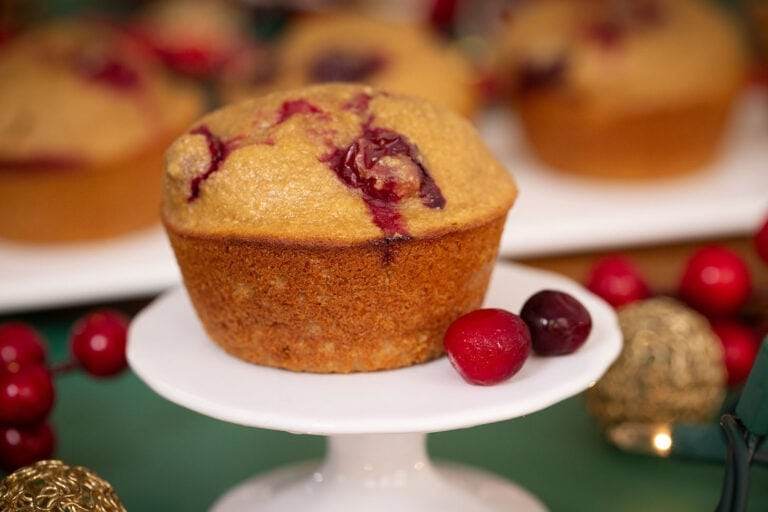 A photo of a Cranberry Orange Blender Muffin on a white plate, on a green table surrounded by holiday decorations.