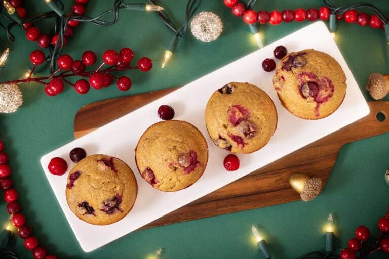 A photo of 4 Cranberry Orange Blender Muffins on a green table surrounded by holiday decorations and cranberries.