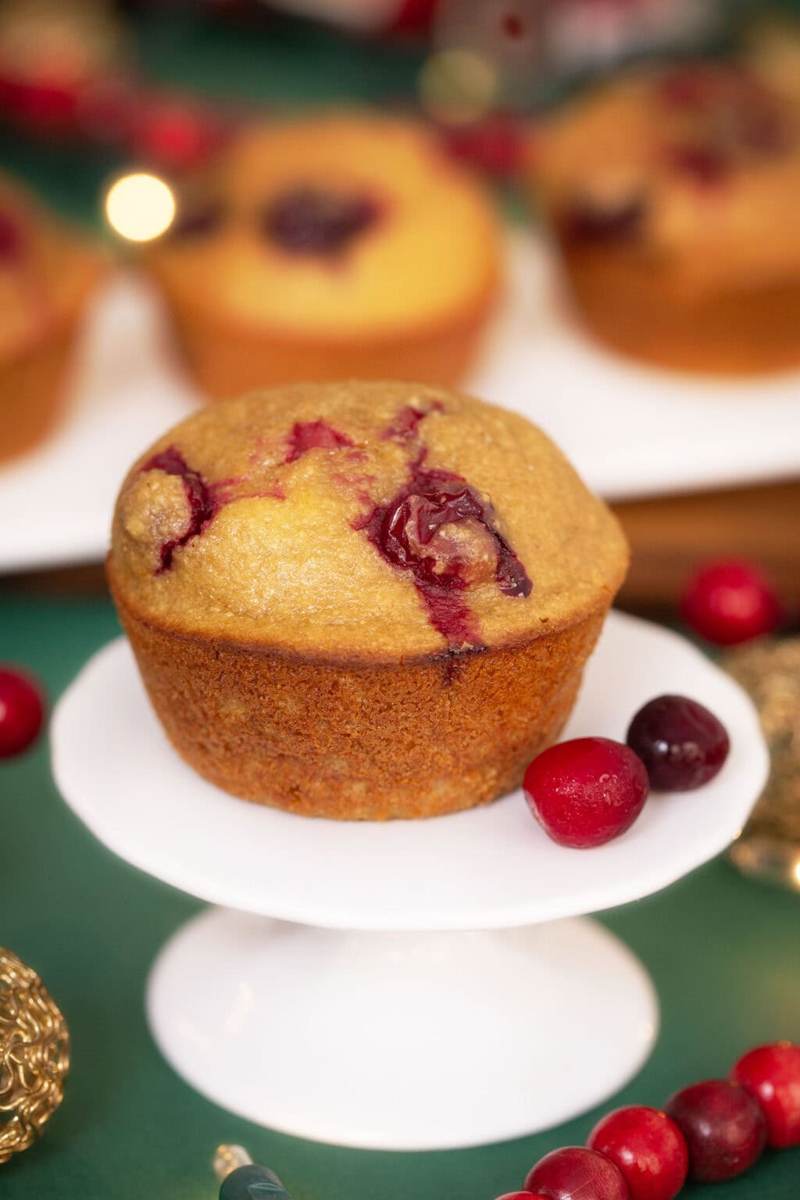 A photo of a Cranberry Orange Blender Muffin on a green table surrounded by cranberries and holiday decorations.