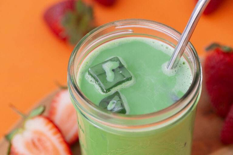 A shot of an iced matcha latte on an orange table, surrounded by fresh strawberries.