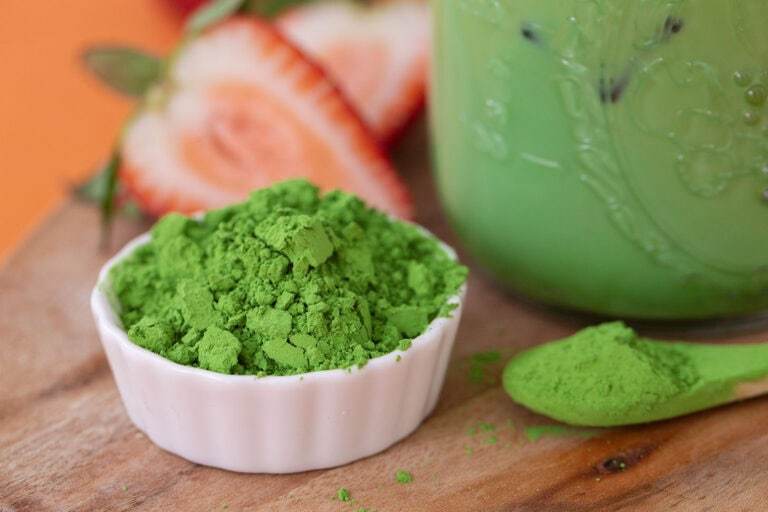 Matcha latte in a glass jar with a bowl of powdered matcha on the side, as well as a mini spoonful of matcha.