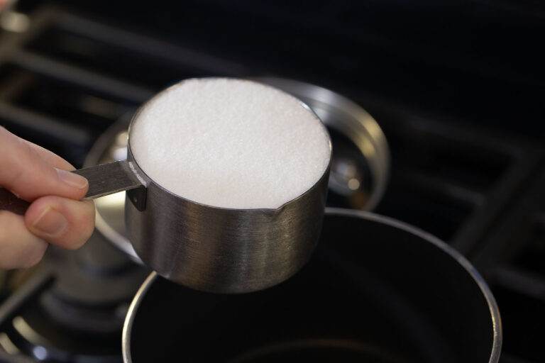 holding a measuring cup filled with sugar over a pot on the stove.