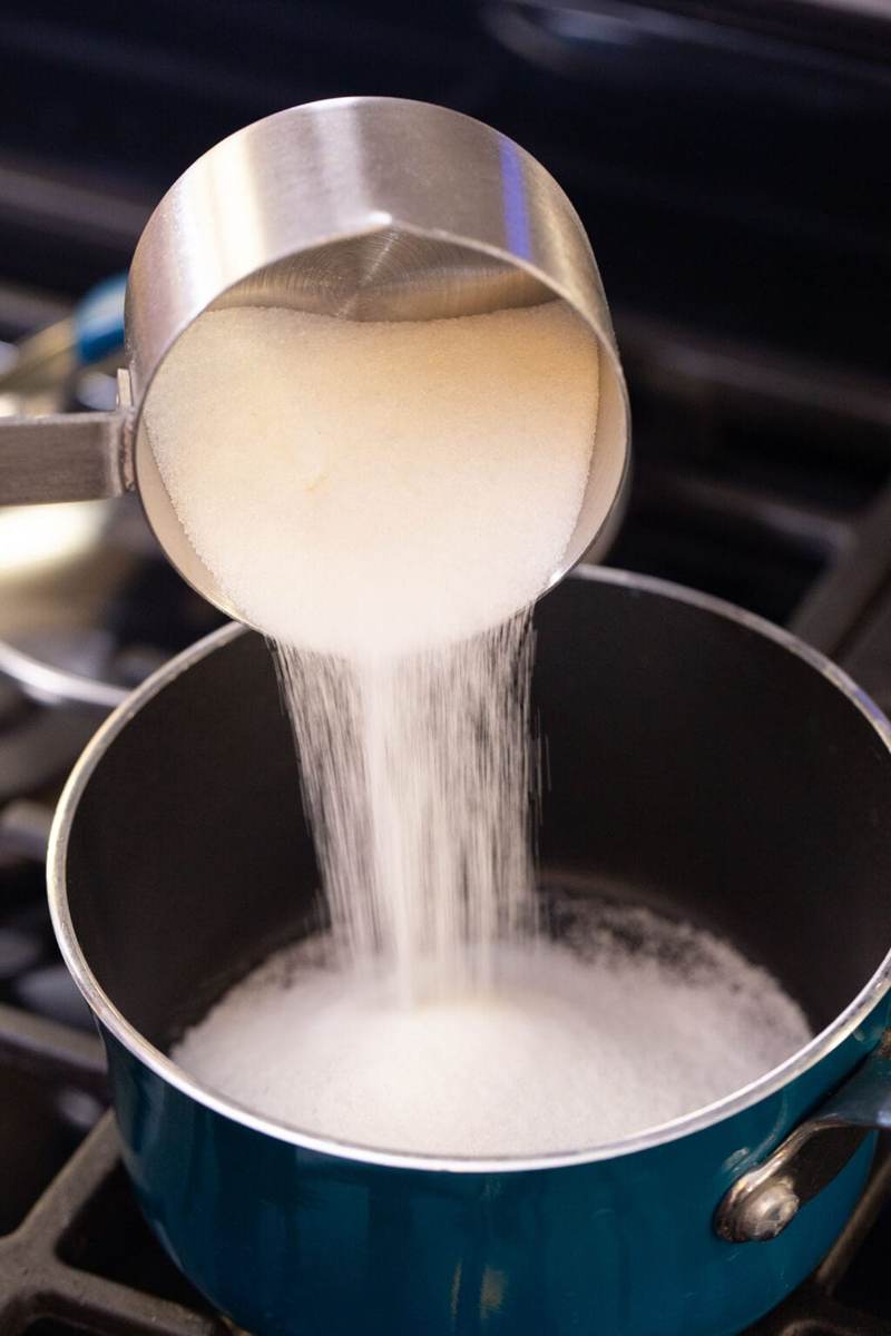 pouring a measuring cup filled with sugar into a pot on the stove.