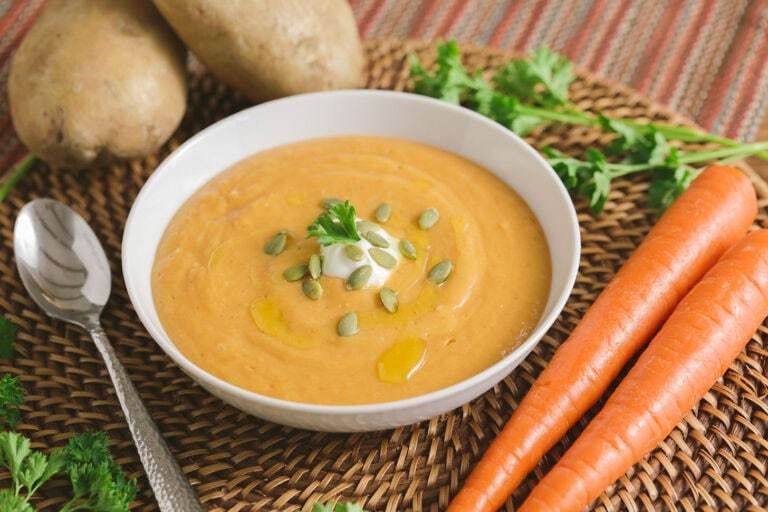 Another photo of the Easy Veggie Soup recipe, wider, surrounded by herbs, carrots, and potatoes. Topped with pumpkin seeds.