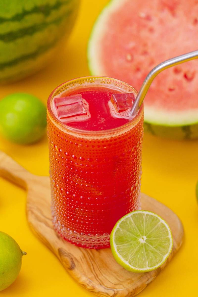 Watermelon agua fresca in a glass, sitting on a cutting board surrounded by ingredients.