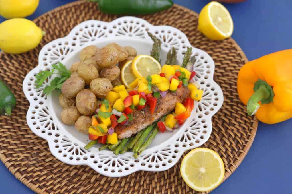 Jerk Salmon on a white plate with Mango Salsa, asparagus, and potatoes. A jalapeno, bell pepper, and lemon on the side.