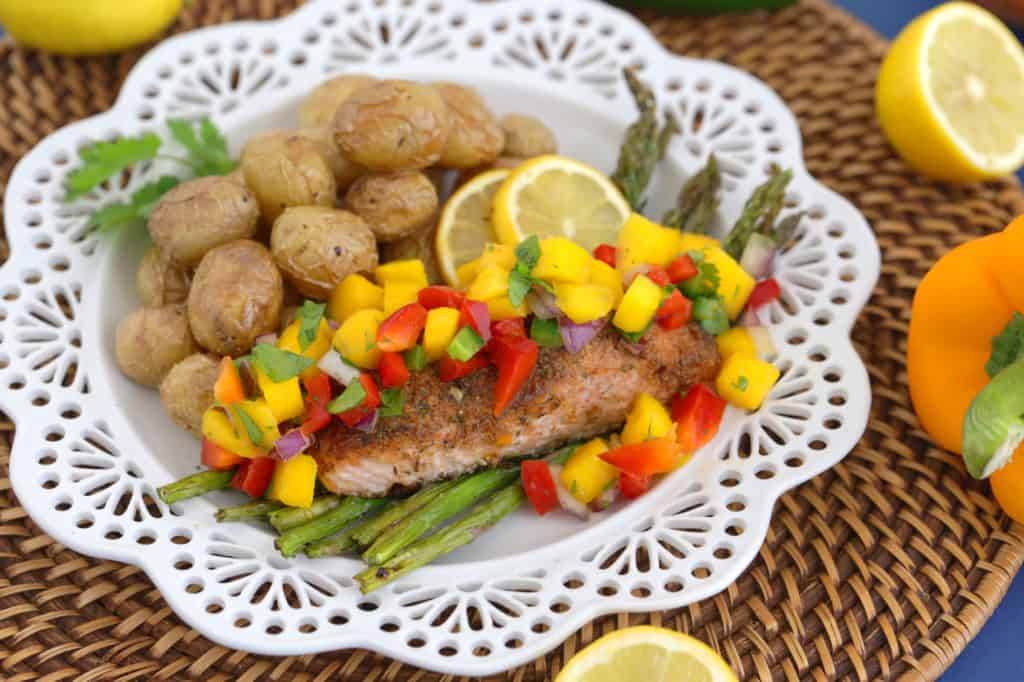 Jerk Salmon on a white plate with Mango Salsa, asparagus, and potatoes. A jalapeno and lemon on the side.