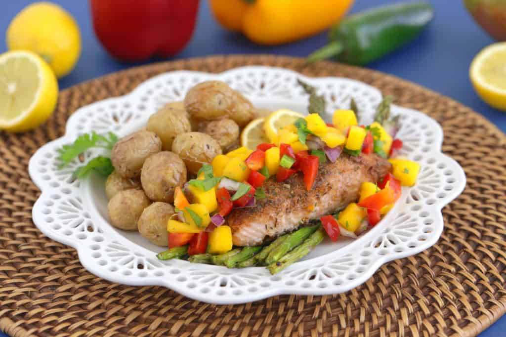 Jerk Salmon on a white plate with Mango Salsa, asparagus, and potatoes. A jalapeno and lemon on the side.