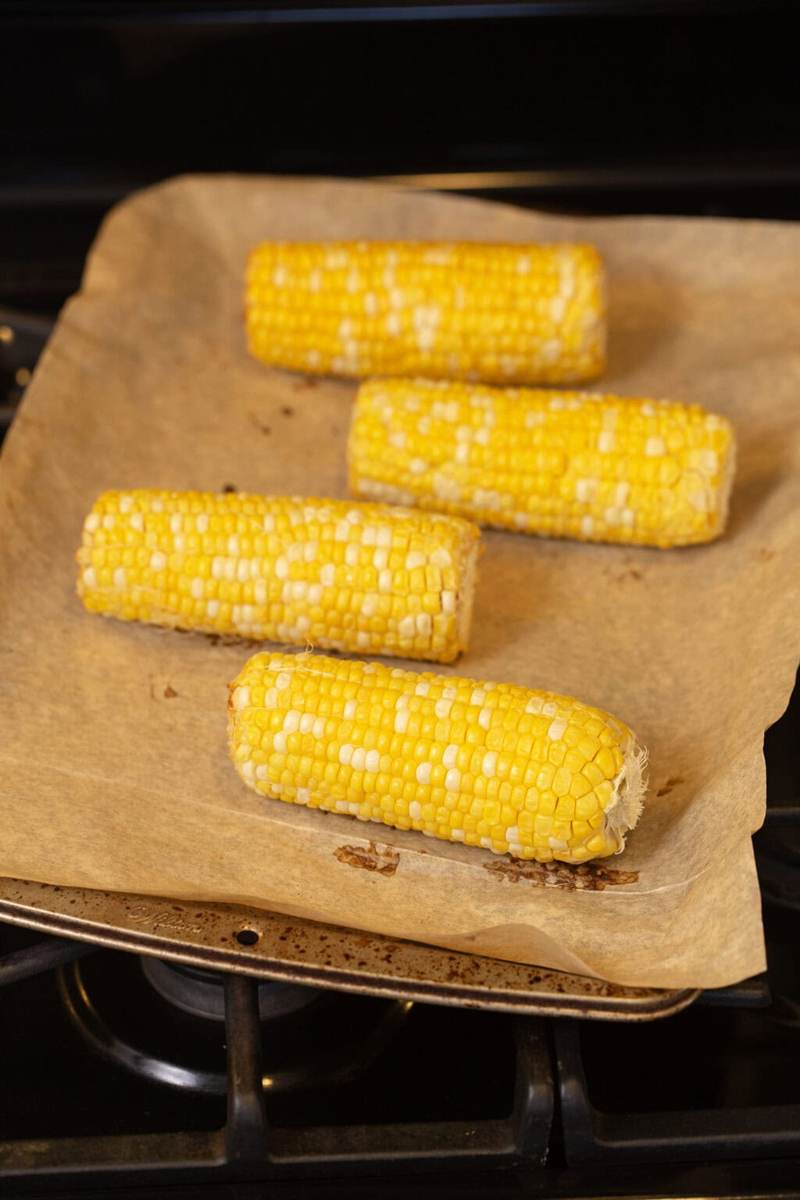 A photo of four corncobs on a baking sheet lined with parchment paper coming out of the oven. They're roasted to perfection.