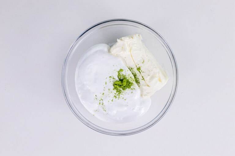 An overhead image of a mixing bowl with marshmallow fluff, cream cheese, and matcha powder.