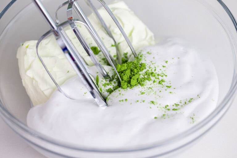 An image of a mixing bowl with marshmallow fluff, cream cheese, and matcha powder, this time with a hand mixer.