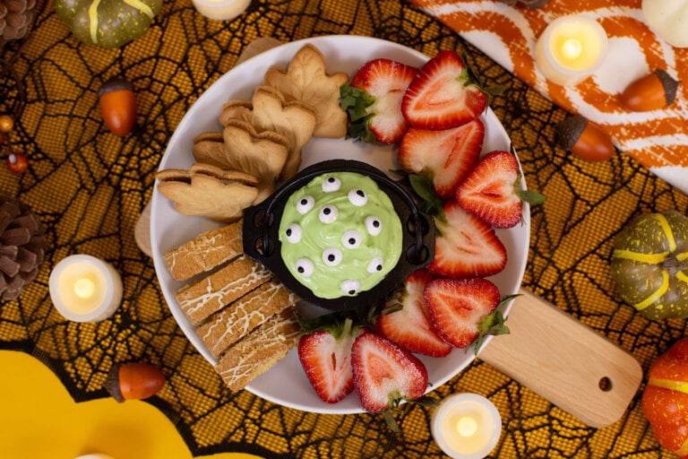 An overhead image of a mini cauldron bowl filled with marshmallow dip covered in candy eyes, sitting on a plate surrounded by cookies and sliced strawberries.