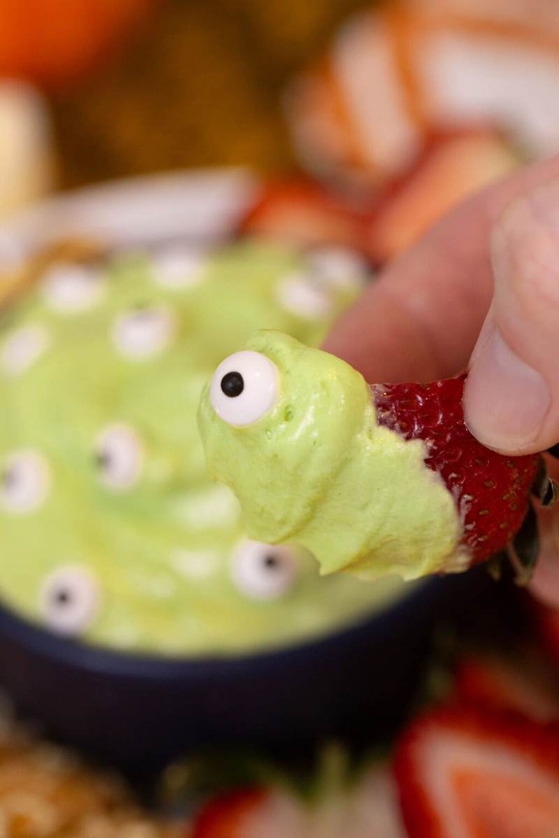 Someone dipping a strawberry slice into the green monster marshmallow dip with a candy eye.