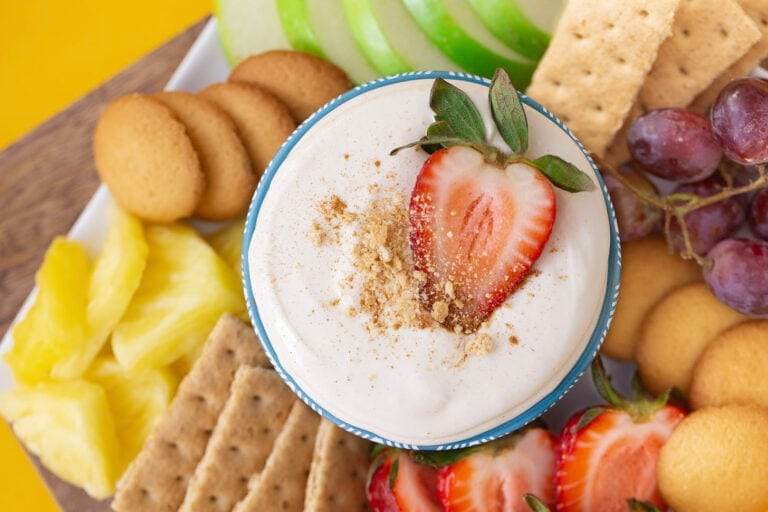 No bake cheesecake dip in a blue bowl, surrounded by dippers such as apples, strawberries, pineapple, and graham crackers.