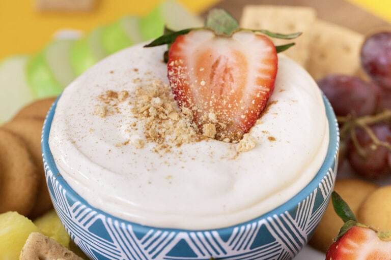 A side view of a blue bowl filled with no bake cheesecake dip, topped with a strawberry, surrounded by various dippers.