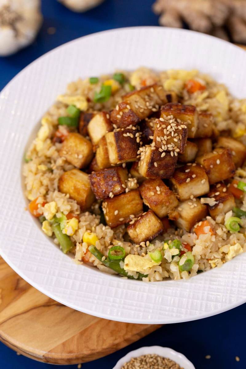 A white bowl on a blue table with cauliflower fried rice, topped with sesame ginger tofu