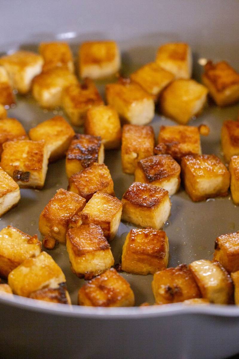 A gray pan on the stove, inside are crispy sesame ginger tofu cubes simmering.