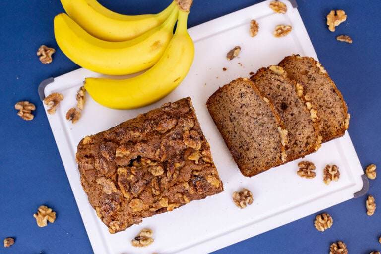 An overhead shot of a loaf of banana bread, half of it cut into slices, on a blue table with a few yellow bananas nearby.