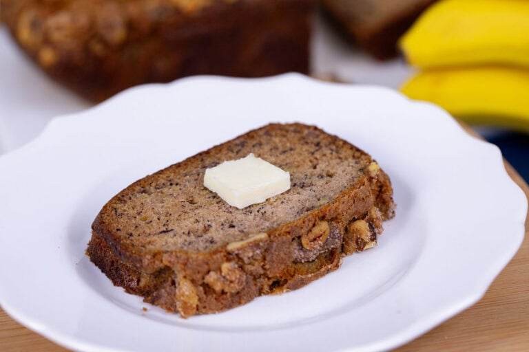 A close up image of a slice of banana bread on a white plate with a pad of butter in the middle.