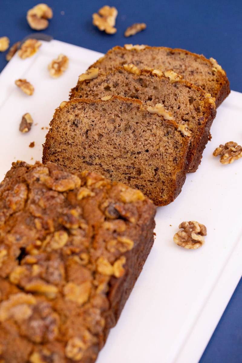 A loaf of banana bread on a white cutting board, half of it cut into slices.