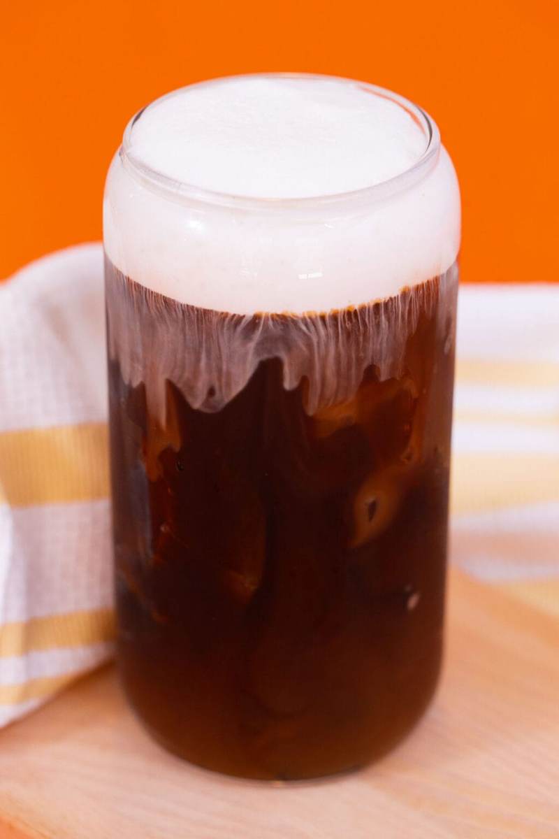 A glass of iced coffee with a vanilla cold foam cap on an orange background.