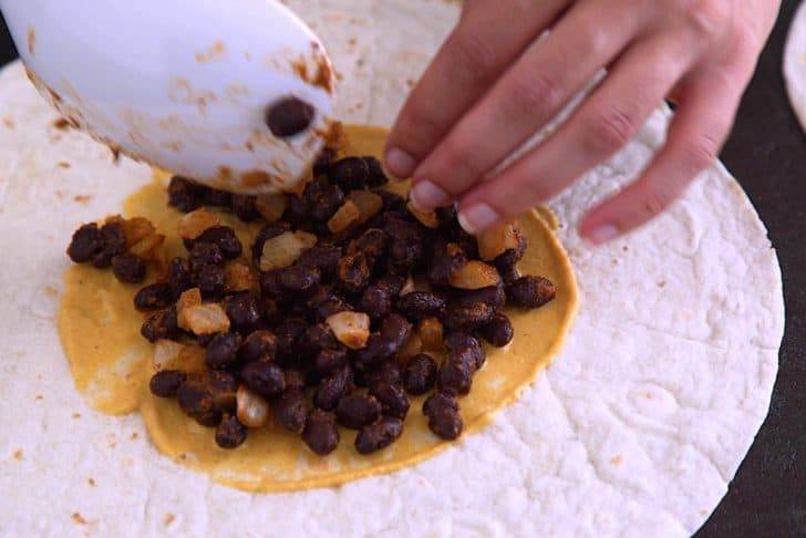 Spread vegan cheese sauce in the center of large tortilla, then add beans and layer on additional fillings.